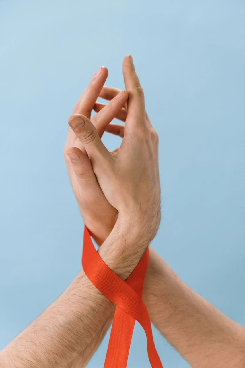 Dos manos unidas con una cinta roja enlazada en las muñecas, sobre un fondo azul claro